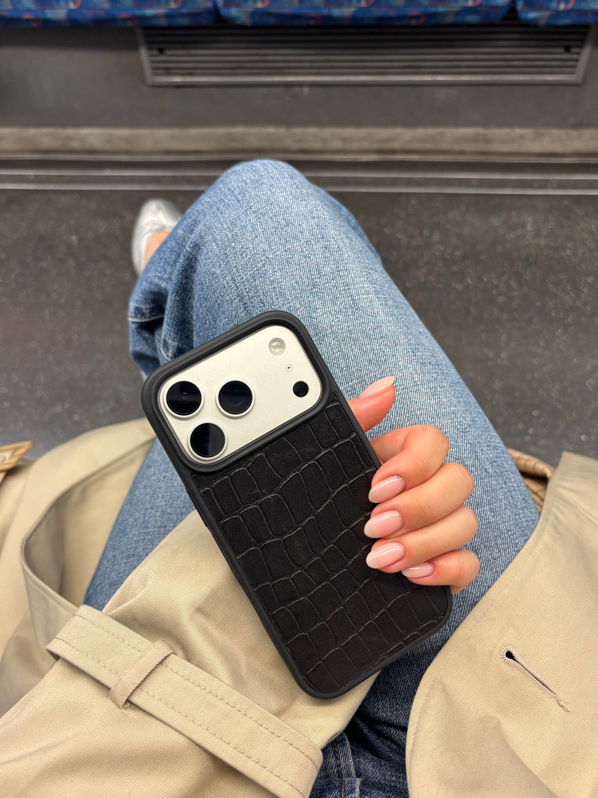 A black phone case with a crocodile-embossed texture, shown on a white smartphone held by a person sitting in a subway or train setting.