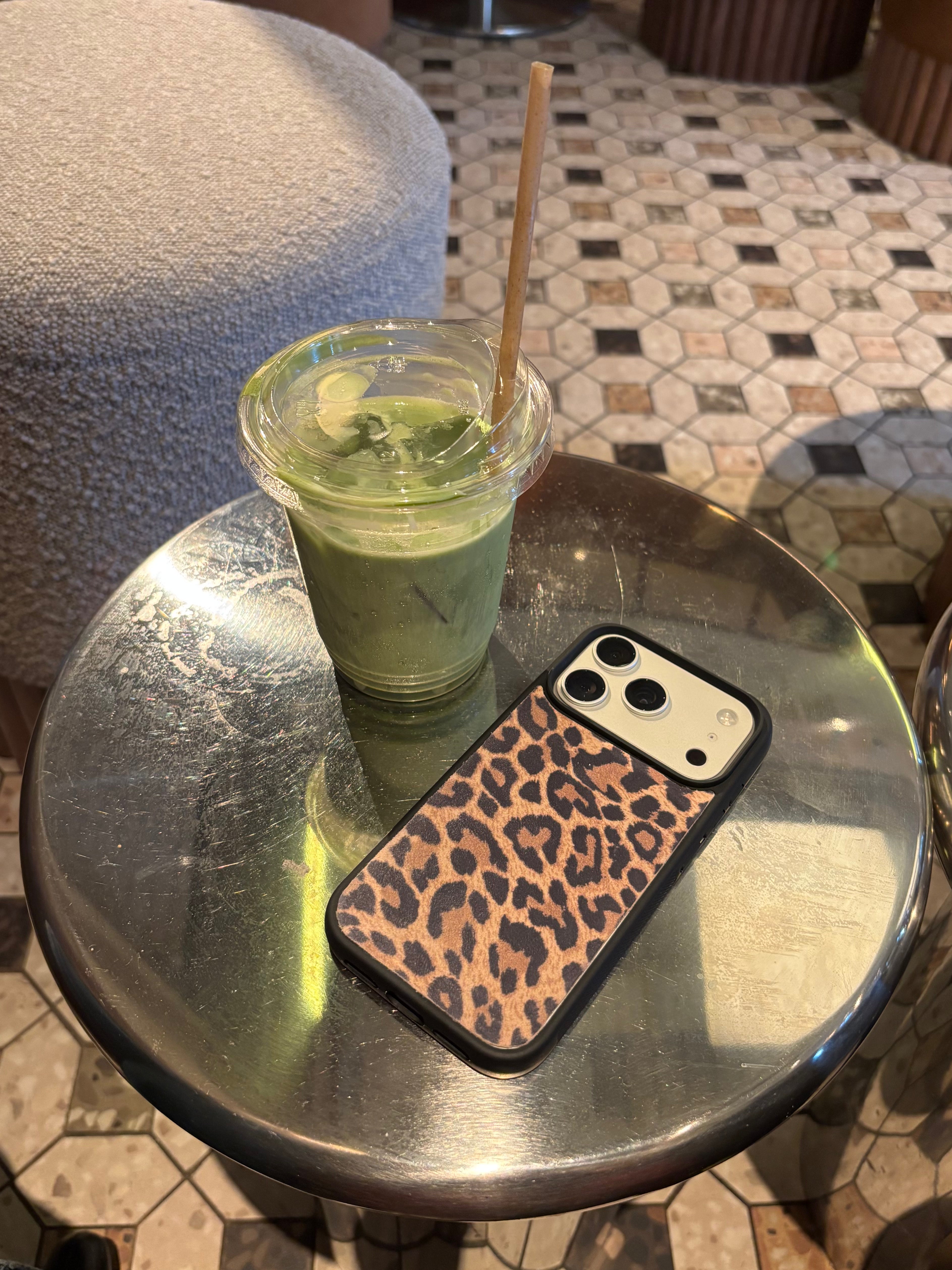 A leopard print phone case on a smartphone and a green iced matcha drink with a straw resting on a round metal table in a cafe setting.