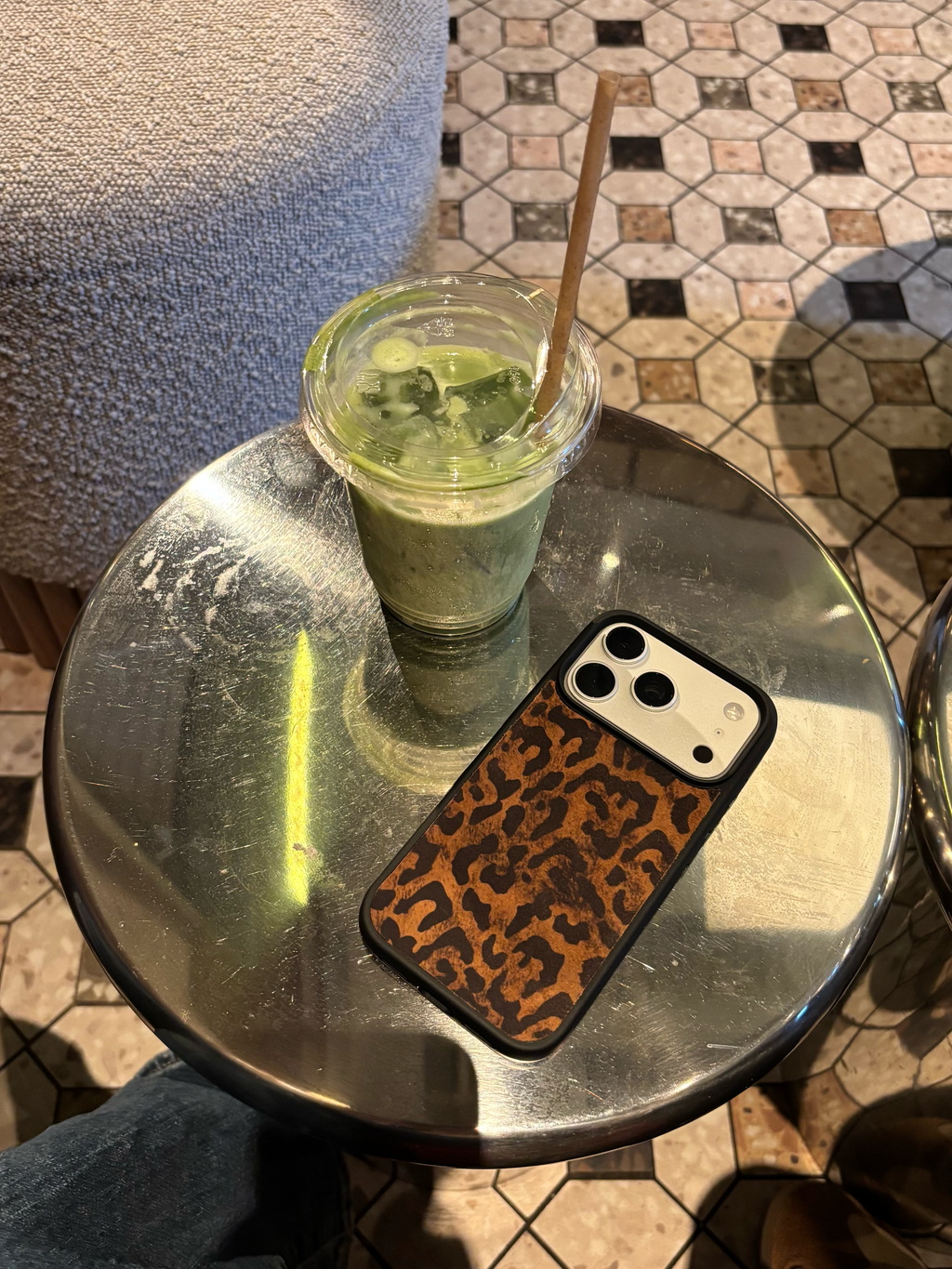 A clear plastic cup of green iced matcha drink with a wooden straw sits on a round metal table next to a smartphone with a brown leopard print case, in a cafe setting with patterned tile flooring.