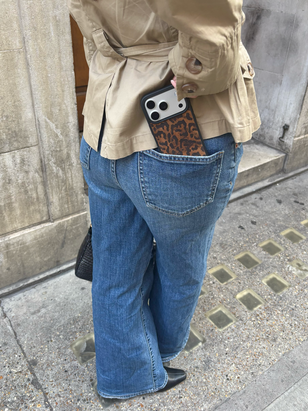 A person wearing a beige belted trench coat and wide-leg blue jeans, shown from the back with a smartphone tucked into the back pocket. The phone has a brown and black leopard print case.
