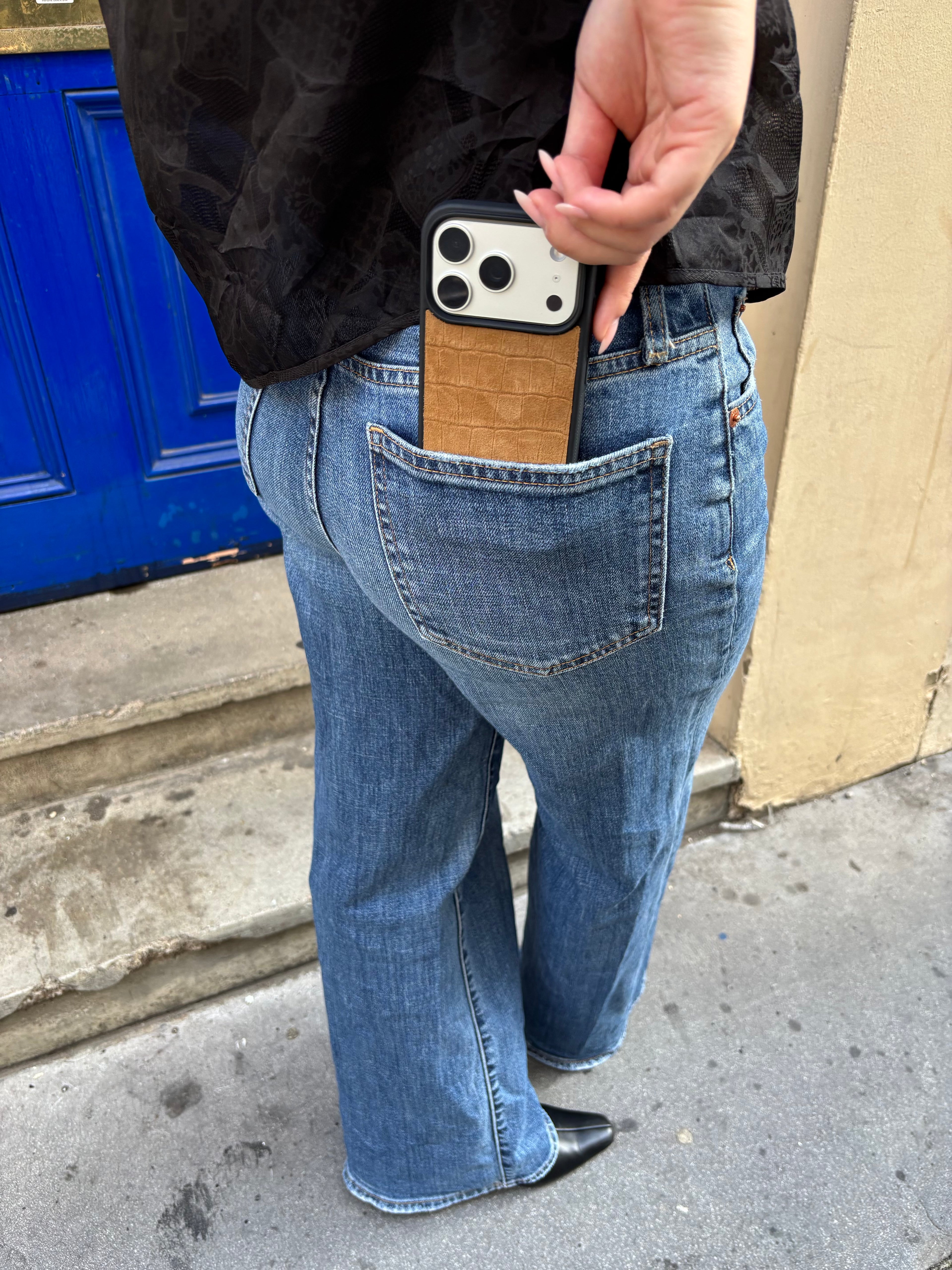 A person wearing blue jeans with a brown leather phone case in the back pocket, holding a black smartphone. The photo is taken outdoors near a blue door and concrete steps.