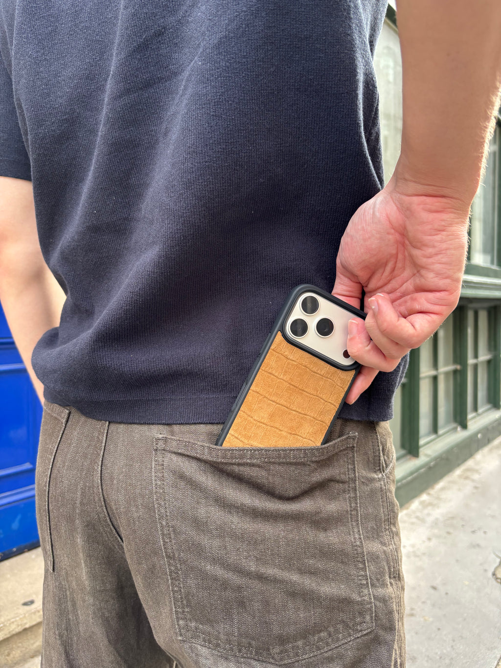 A close-up lifestyle shot of a person wearing a navy blue t-shirt and brown pants, showing a phone with a tan textured leather case tucked into the back pocket.