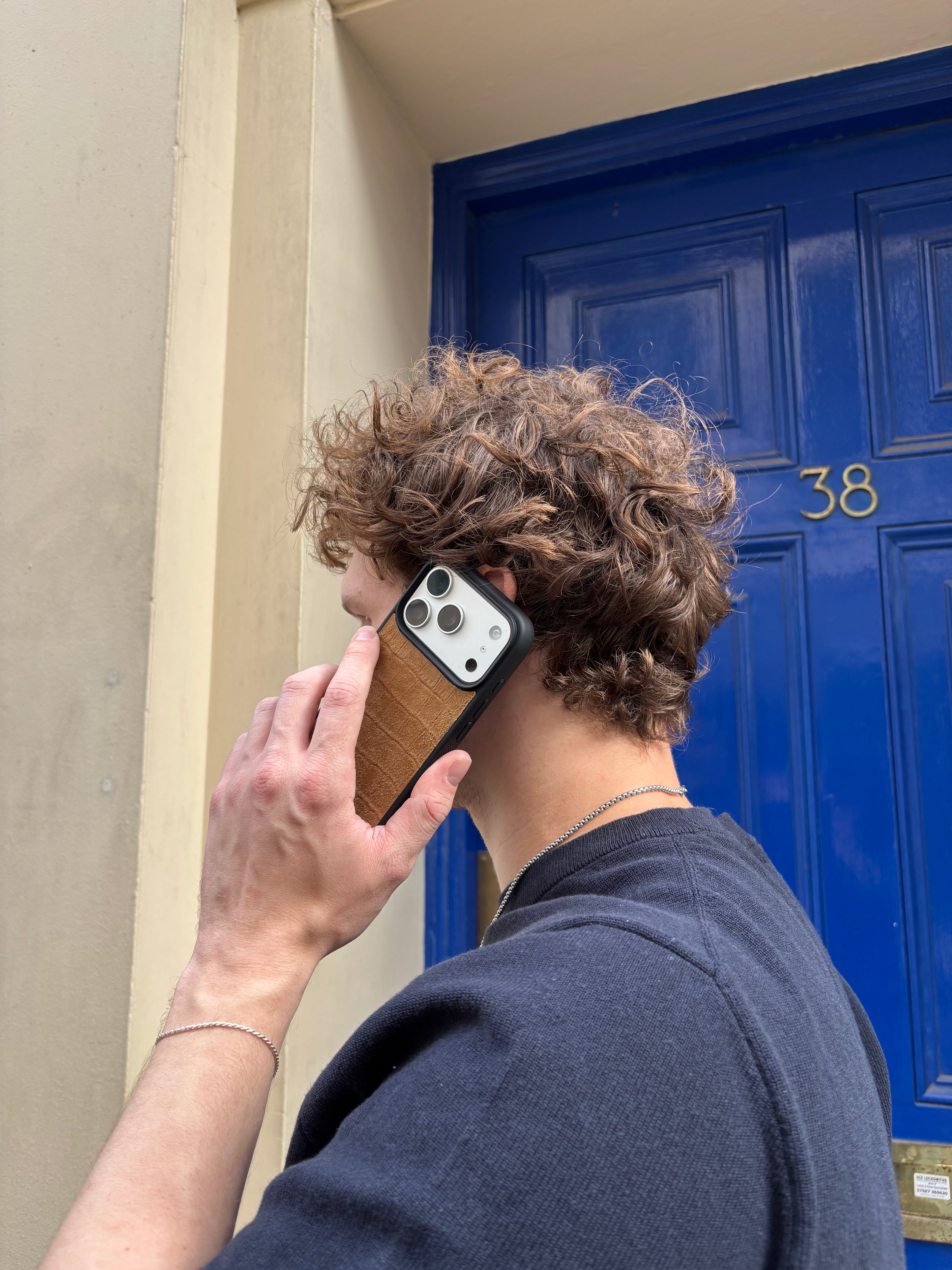 A person with curly brown hair wearing a dark navy t-shirt and holding a smartphone with a brown wooden-textured case next to a bright blue door.