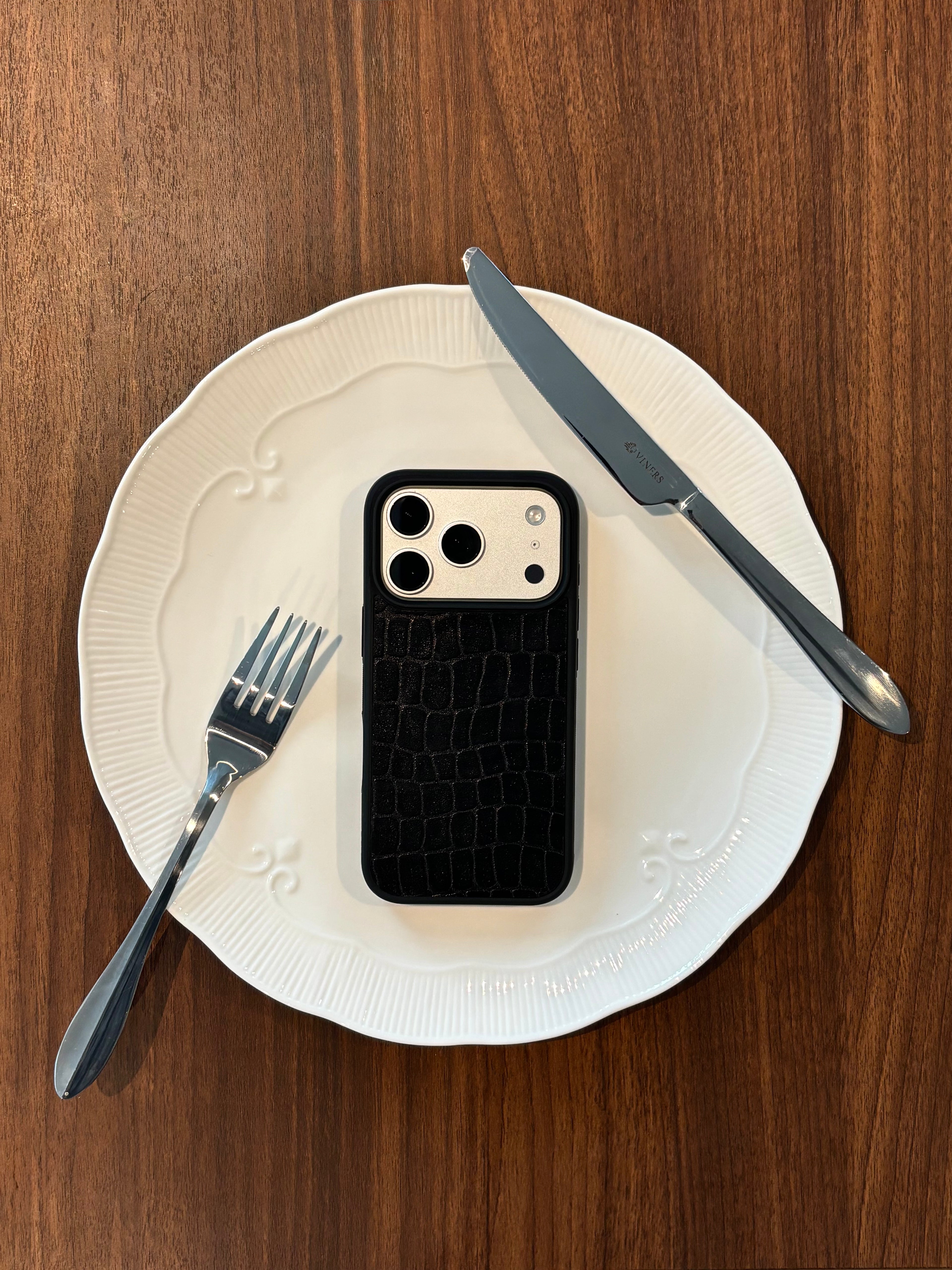 A black crocodile-embossed phone case on a smartphone, styled on a white plate with a knife and fork on a wooden table.