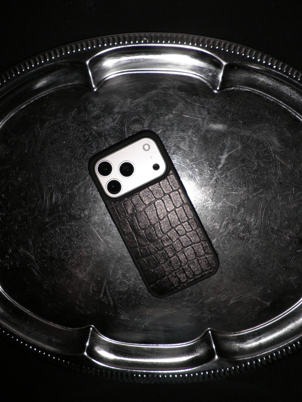 A black textured phone case with a crocodile skin pattern, shown on a silver metal tray against a black background.