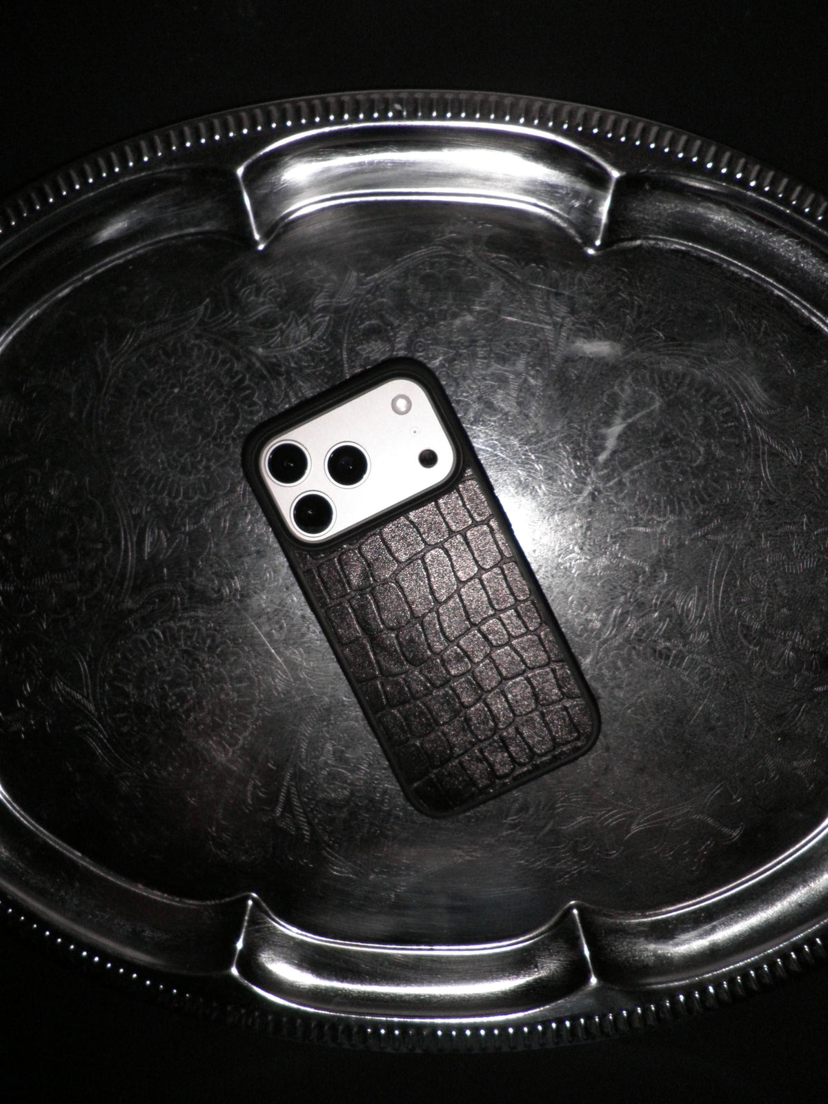 A black textured phone case with a crocodile skin pattern, shown on a silver metal tray against a black background.