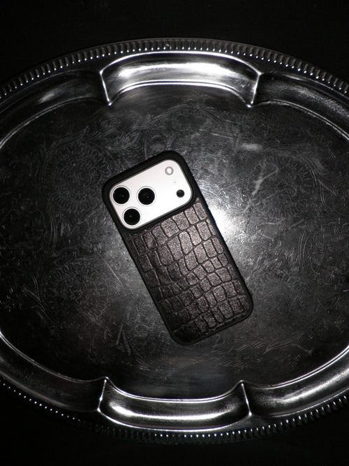 A black textured phone case with a crocodile skin pattern, shown on a silver metal tray against a black background.