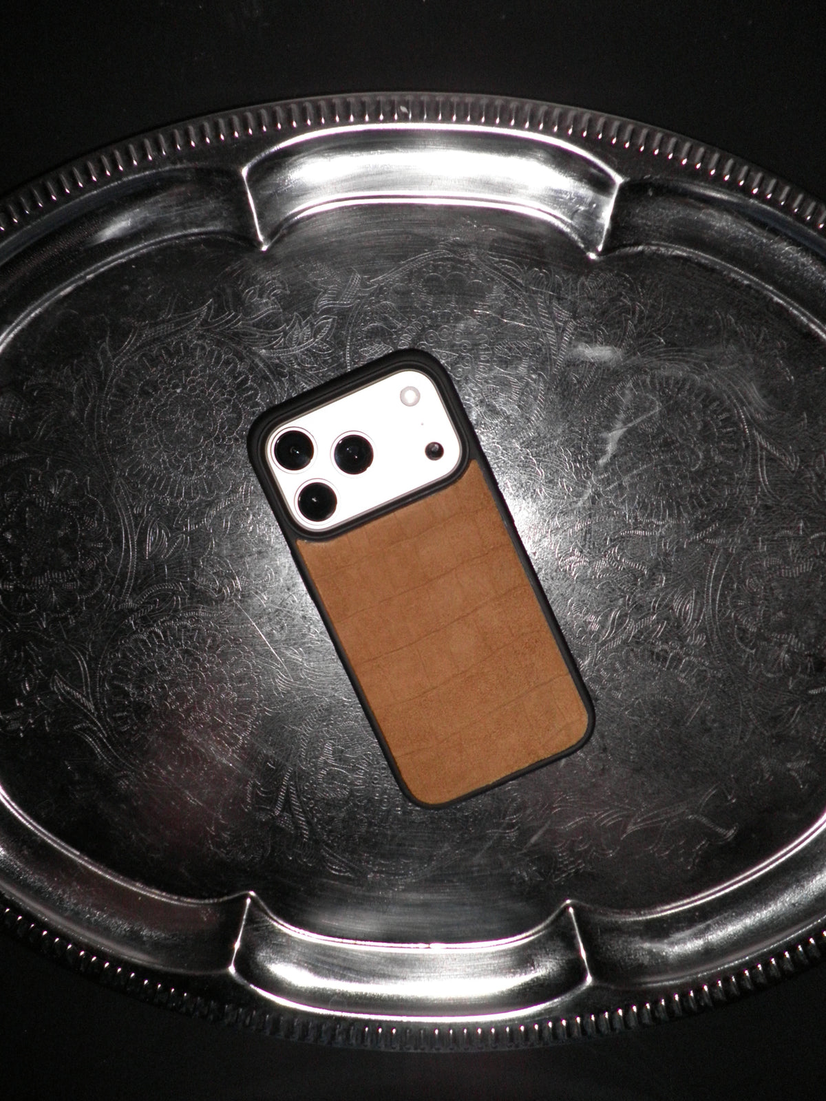 A phone case with a brown textured leather back and black border, shown on a smartphone resting on a reflective silver metal tray.