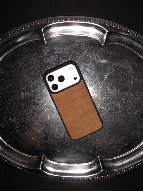 A phone case with a brown textured leather back and black border, shown on a smartphone resting on a reflective silver metal tray.