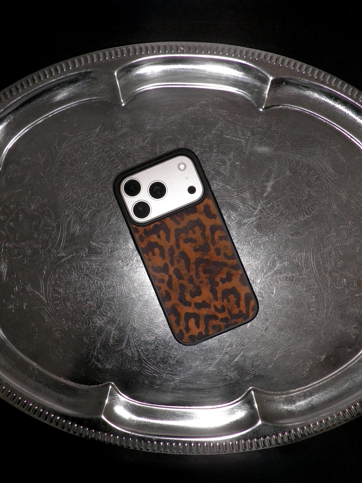 A smartphone case with a brown and black leopard print pattern, shown on a silver metal tray against a black background.